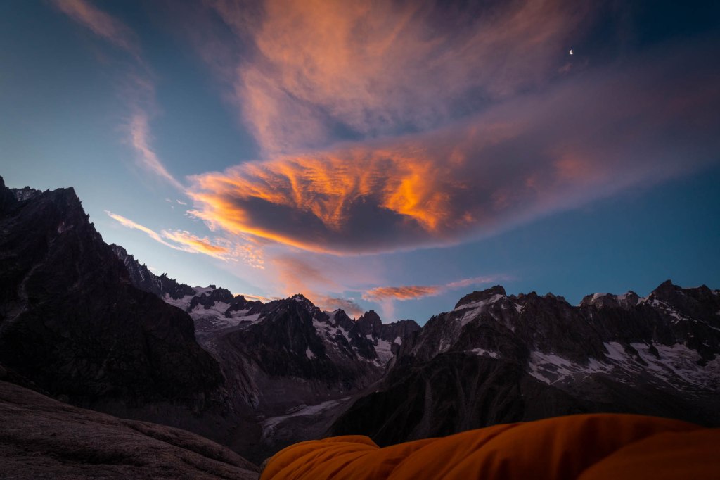 A breathtaking view of the French Alps at sunset, showcasing dramatic mountain peaks and vibrant clouds illuminated by orange hues, with a hint of a moon in the sky.