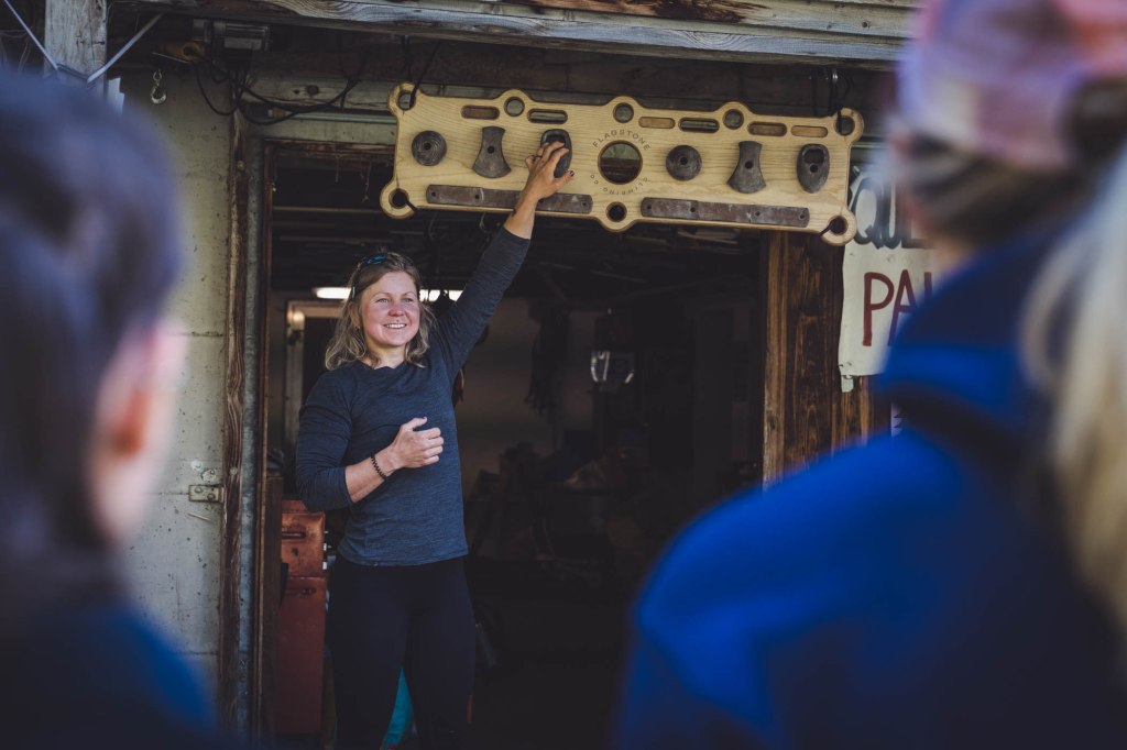 A woman demonstrating climbing equipment in a workshop, smiling and engaging with the audience, with a climbing wall and gear displayed in the background.