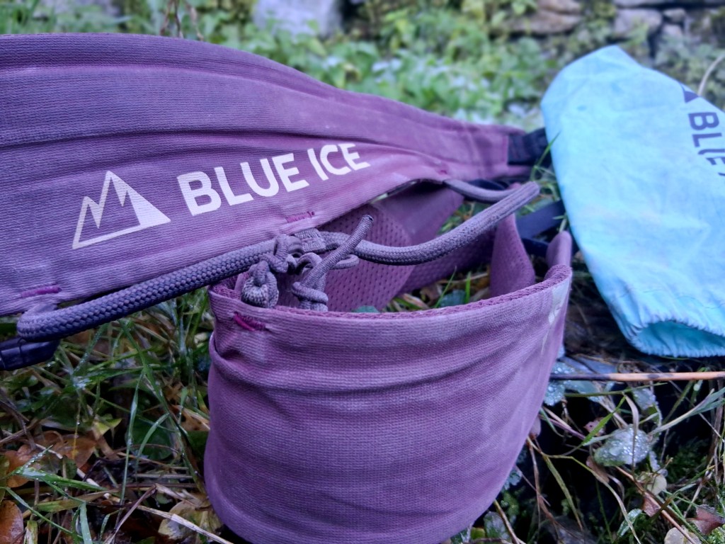 A close-up shot of Blue Ice climbing gear, including a purple adjustable harness and a light blue gear pouch, placed on grassy ground with natural elements in the background.